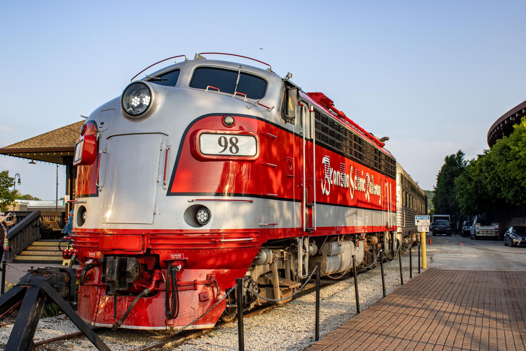 A red and silver locomotive marked with the number "98" sits on display at a station. It is part of the Branson Scenic Railway, with clear skies in the background and surrounding station details visible.